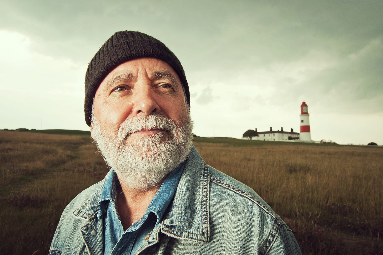 Man with grey beard standing down the hill from a lighthouse