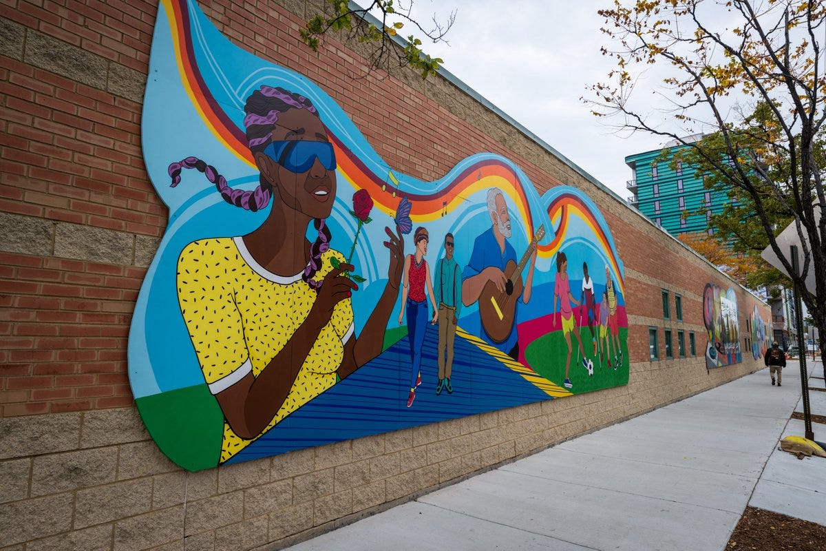 Photo is of a mural created by Artist Sam Kirk. The mural has a vibrant blue background with individuals of diverse disabilities including a woman with braids wearing glasses, smelling a rose with a butterfly on her finger, a couple holding hands with one wearing glass. Next to them is a man with gray hair and facial hair playing the guitar, and right of him are four young people playing soccer. This mural is located on the building of the Central West Community Center