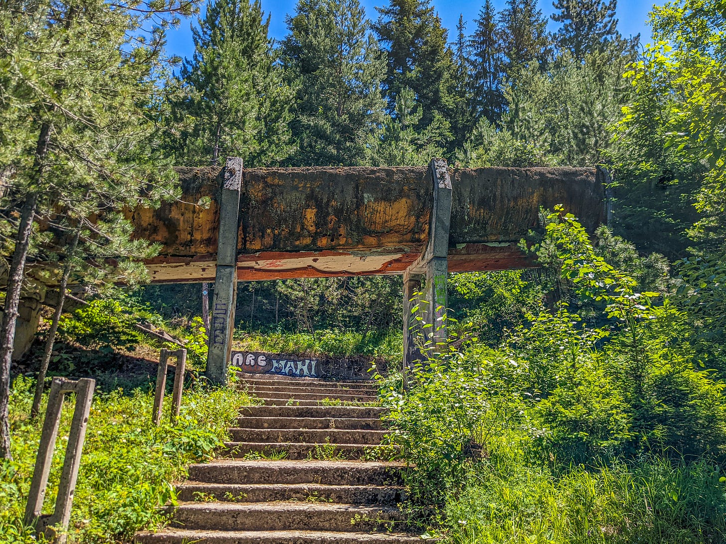 A ruined portion of the Olympic bobsleigh run in front of a backdrop of trees. The concrete run supported by two pillars is stained black and orange. 
