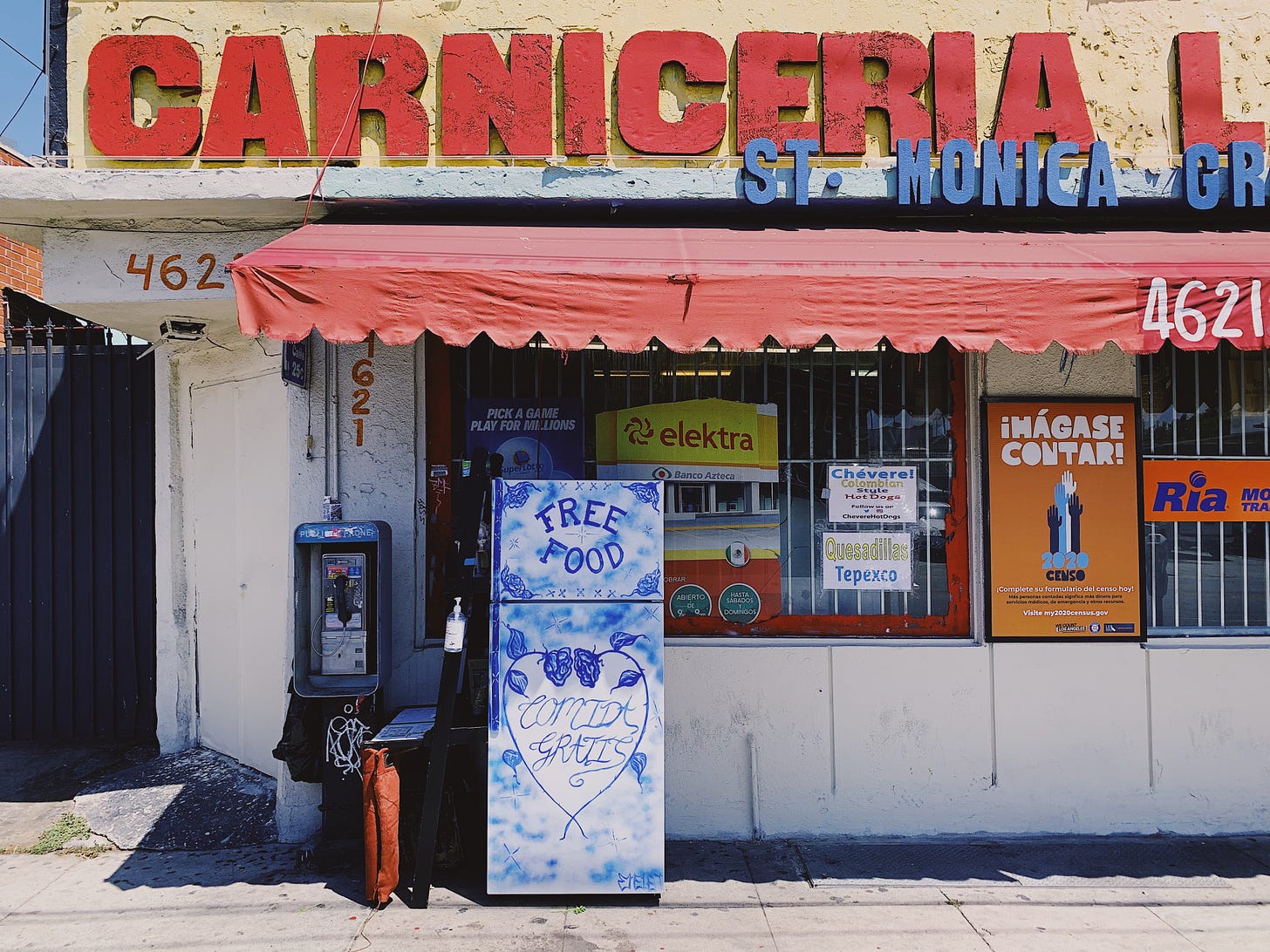 Yellow and white building with the words “Carniceria” in red block letters at the top and a red awning below. A white fridge with blue paint and the words “free food, comida gratis” written on the front. The fridge is next to a payphone.