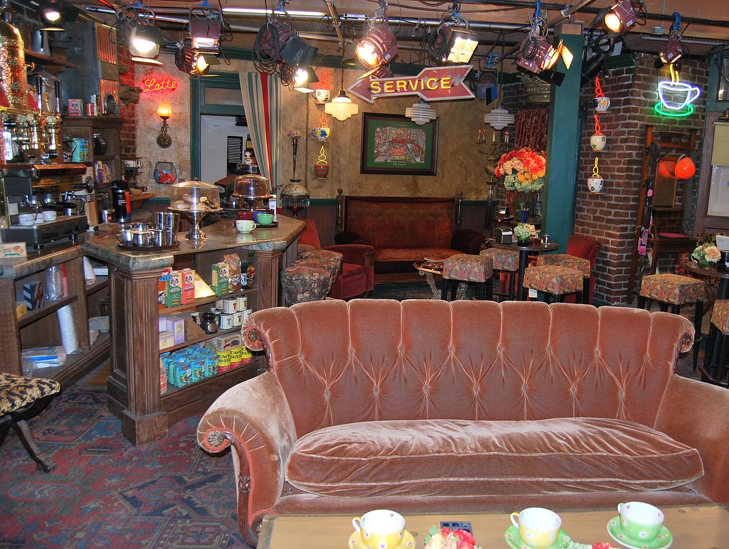 A photo of the Central Perk Coffee Shop set from the TV show friends. A worn, vintage-style red/orange couch dominates the foreground with a cluttered coffee county and stools in the background.