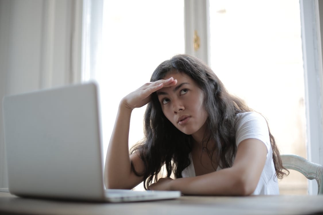 Irritated ethnic female entrepreneur in casual wear sitting at table with netbook and touching head while waiting for internet connection during remote work