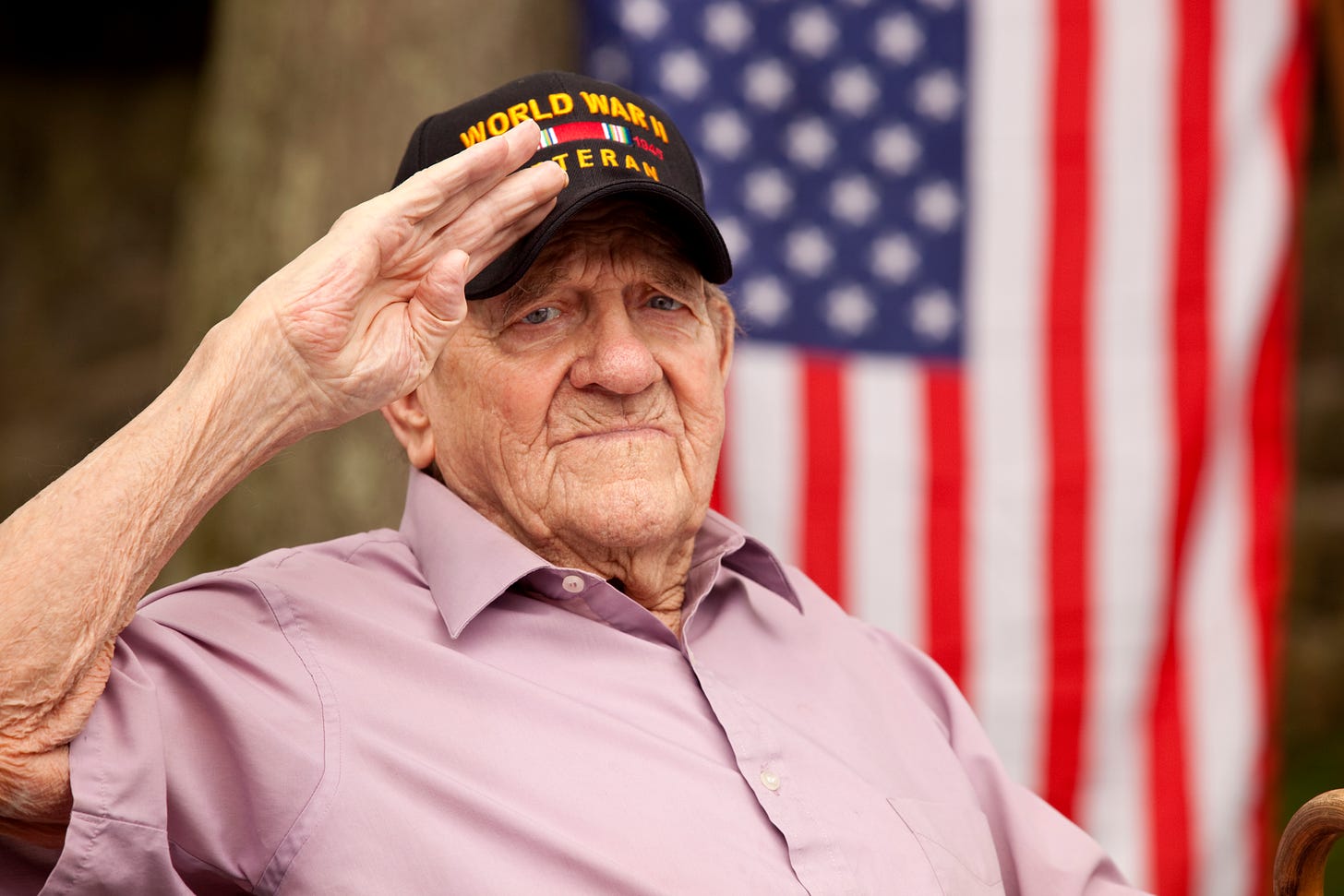 World War II vet in baseball cap saluting in front of vertical American Flag