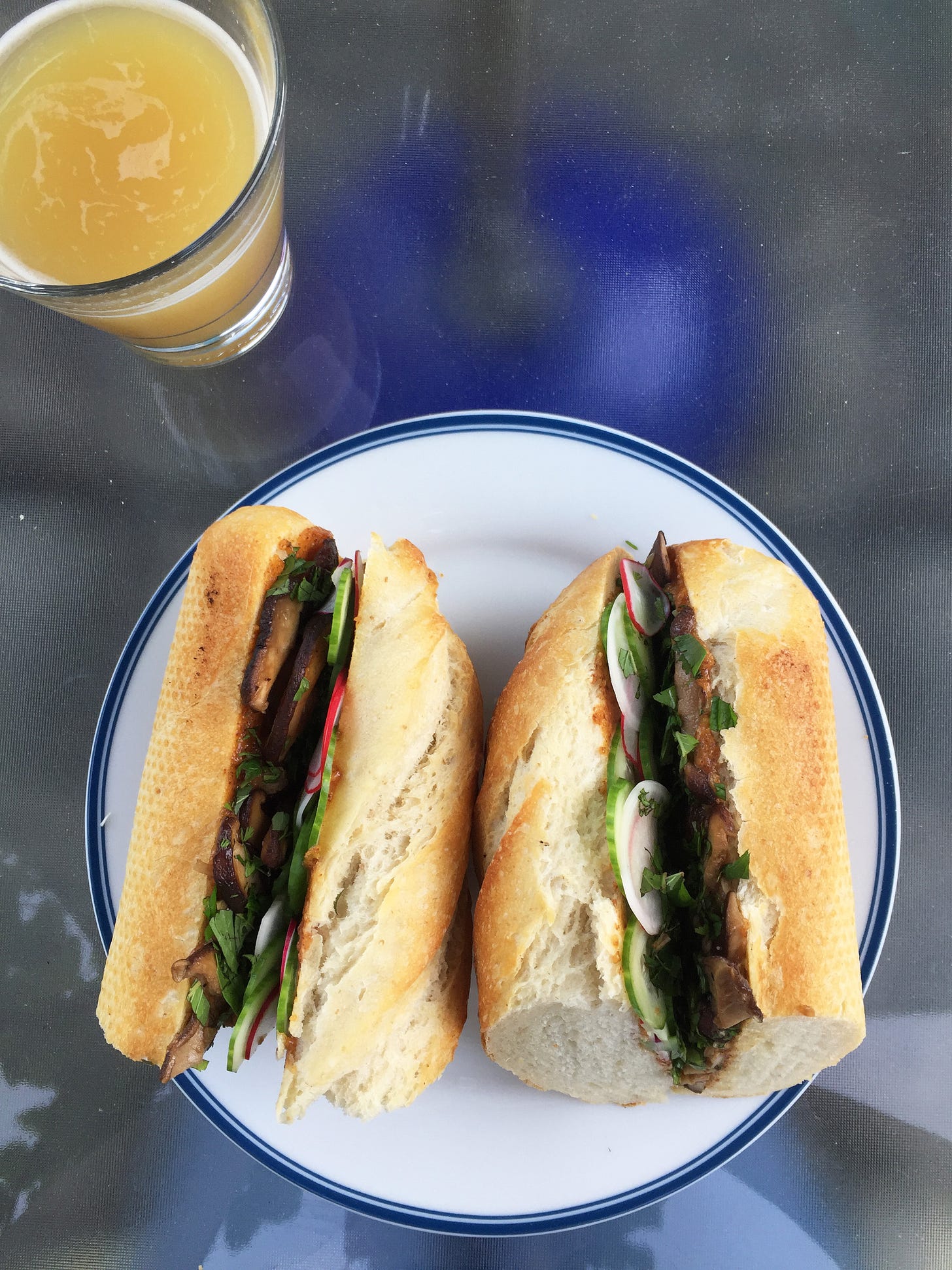 from above, a baguette sandwich cut in half with herbs, radish, cucumber, and mushrooms visible. In the upper left corner sits a cloudy glass of IPA.