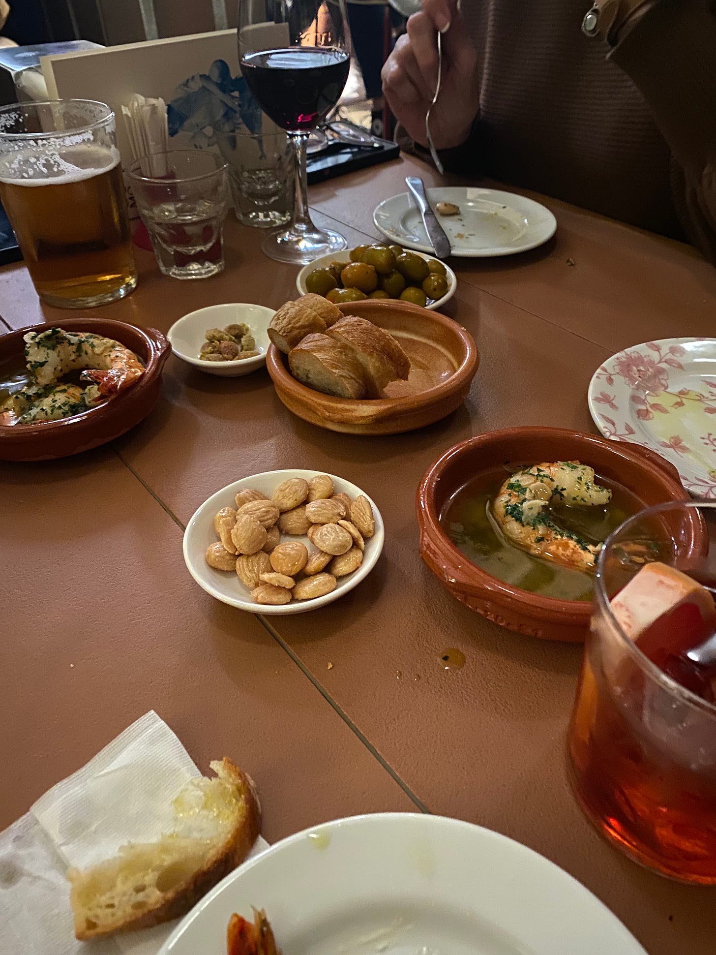 a restaurant table with a scattering of small dishes and plates, partially eaten. A cocktail peeks out of the lower right corner next to a plate and a crust of bread; in the centre are a dish of marcona almonds and two dishes of prawns in garlic sauce, a dish with two slices of baguette, and behind it, a white bowl of marinated olives next to a ramekin of their pits.