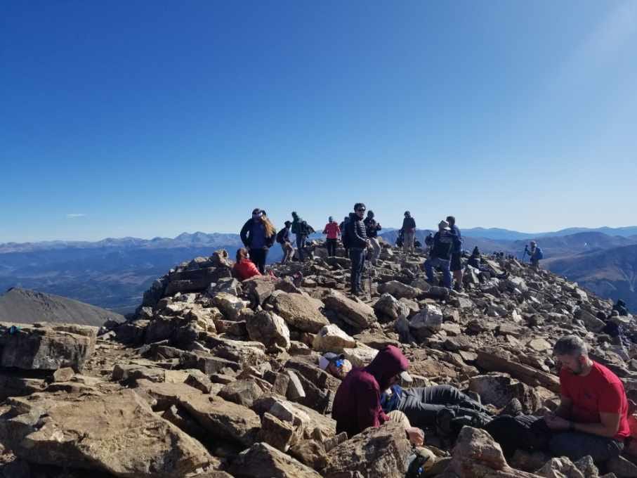 a busy crowd packs onto Quandary peak. The bright blue sky dominates most of the frame. Clusters of people wait on the rocky summit for photos.