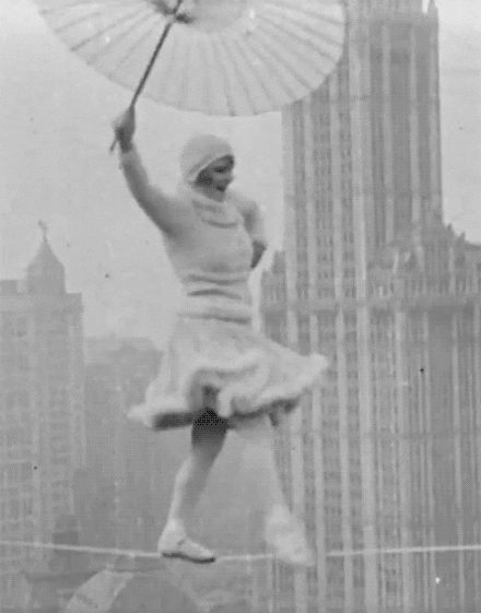 GIF. Black and white 1930s New York City scene of a woman dancing and balancing on a tightrope while holding an umbrella over her head. She’s wearing a nice-looking beanie and a fur-trimmed knee-length skirt.