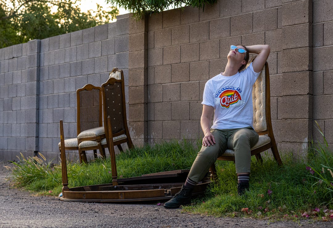 A young woman sitting in front of a concrete block wall on an abandoned-looking wooden dining room chair, with a couple more chairs and a broken table lying nearby. She is wearing the Today in Tabs “You Can Always Quit” t-shirt in white, with its cheerful rainbow logo, and she gazes up at the sky in sunglasses, thinking about the much better future ahead now that she’s quit.