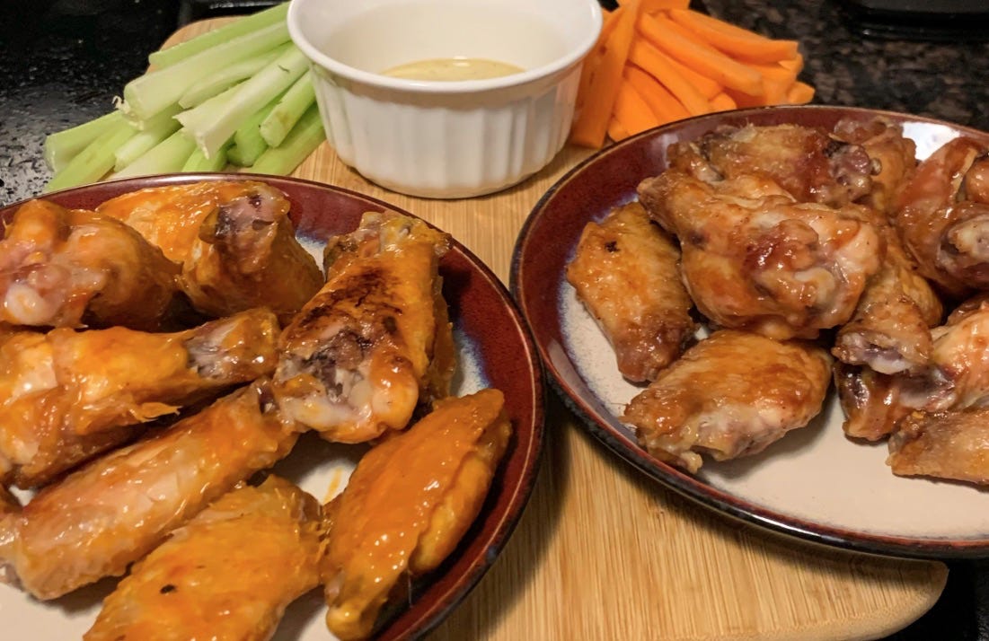 Image shows two plates of wings, with two different sauces, on top of a wooden board with chopped celery and carrot sticks, and a ramekin of a creamy sauce.