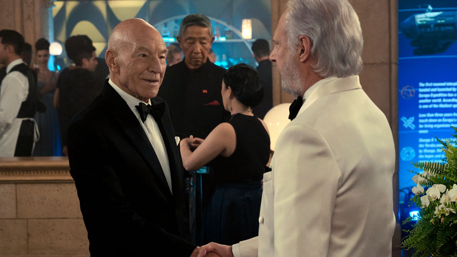 This image shows Jean-Luc Picard (played by Patrick Stewart, left) being greeted by Adam Soong (played by Brent Spiner, right) at the Europa Mission gala. Picard wears a traditional black tuxedo, while Adam wears a white tux. This image shows Jean-Luc Picard (played by Patrick Stewart, left) being greeted by Adam Soong (played by Brent Spiner, right) at the Europa Mission gala. Picard wears a traditional black tuxedo, while Adam wears a white tux.