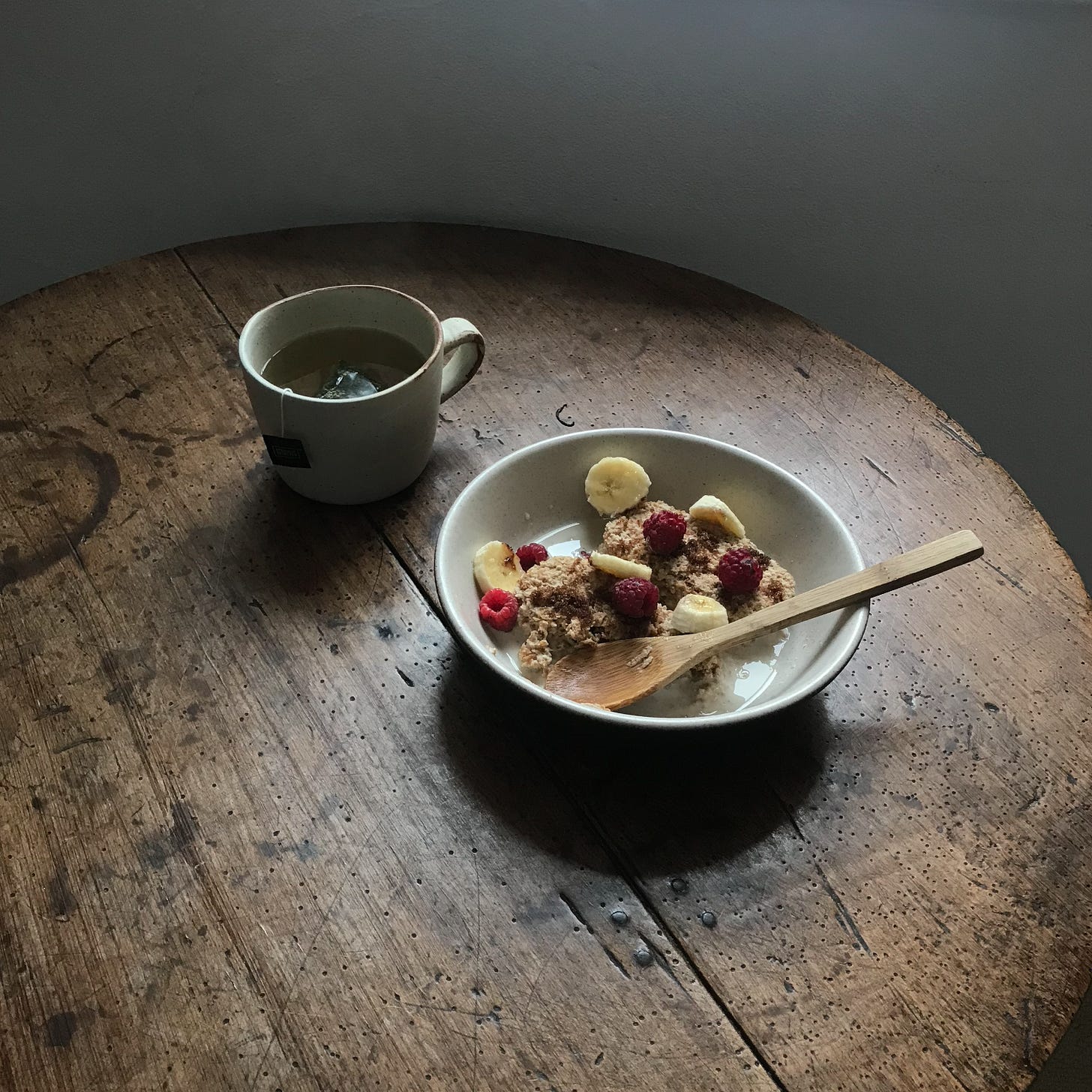 A bowl of weetabix with raspberries and banana, on a wooden table beside a mug of tea. 