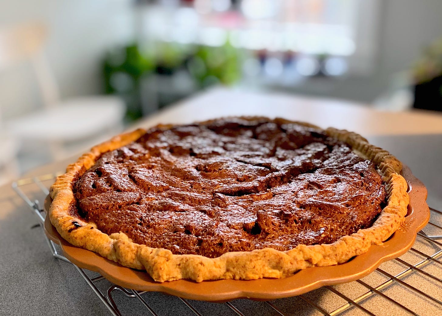A bourbon and chocolate pecan pie rests in a copper-colored pie tin on a cooling rack in a kitchen. 