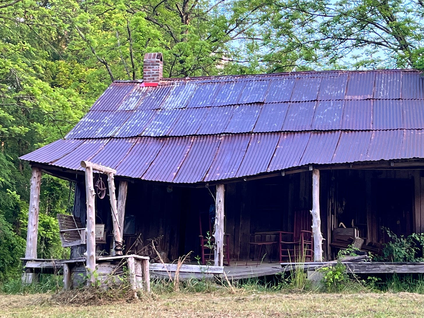 Image of an old shack on the side of the road, with a dropping rusted corrugated steel roof, and several old chairs in the shadows.