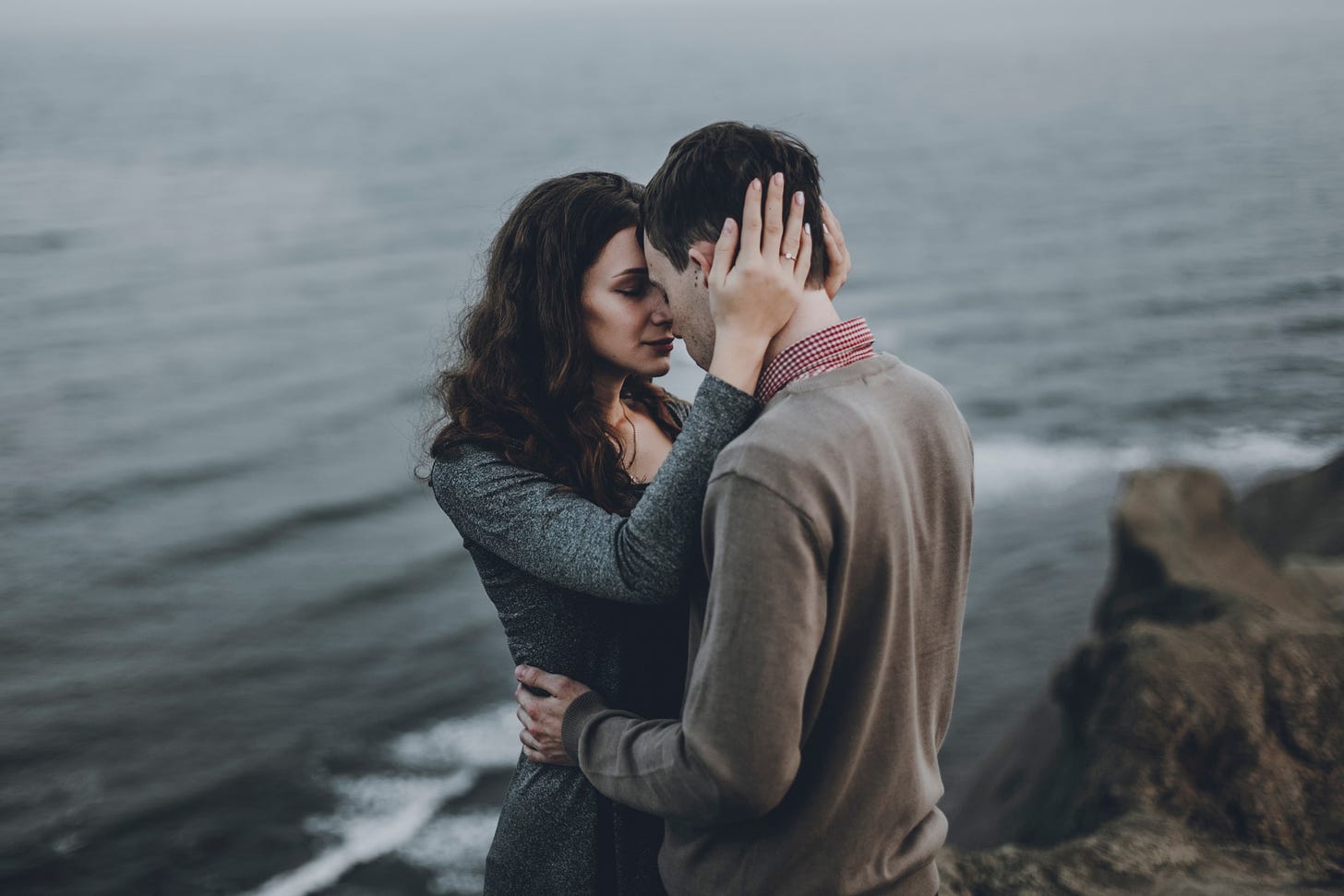 A couple embraces on the edge of a cliff on a cloudy day. The ocean moves behind them.