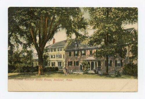 postcard image of two houses. House in the foreground is built of stone with dark window shutters and a gazebo-shaped porch over the central front door. The front yard is shaded by tall oaks.