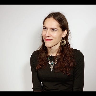 Photo of Alyssa Gonzalez. She is a woman with wavy brown hair and brown eyes, smiling just past the camera, wearing a black shirt, necklace, and large gold earring, in front of a gray background.