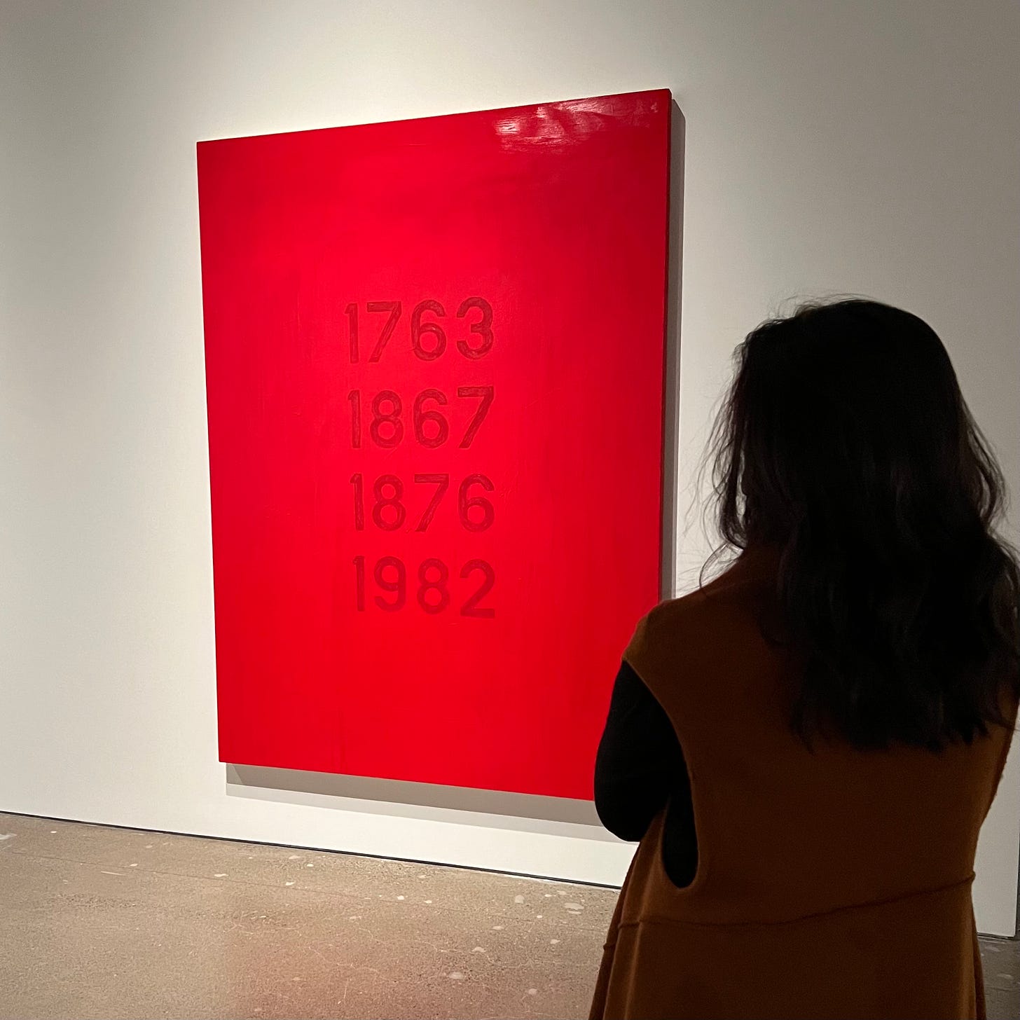 The back of a woman standing to the right side and in front of a painting by Robert Houle. The painting is red with four years on it.