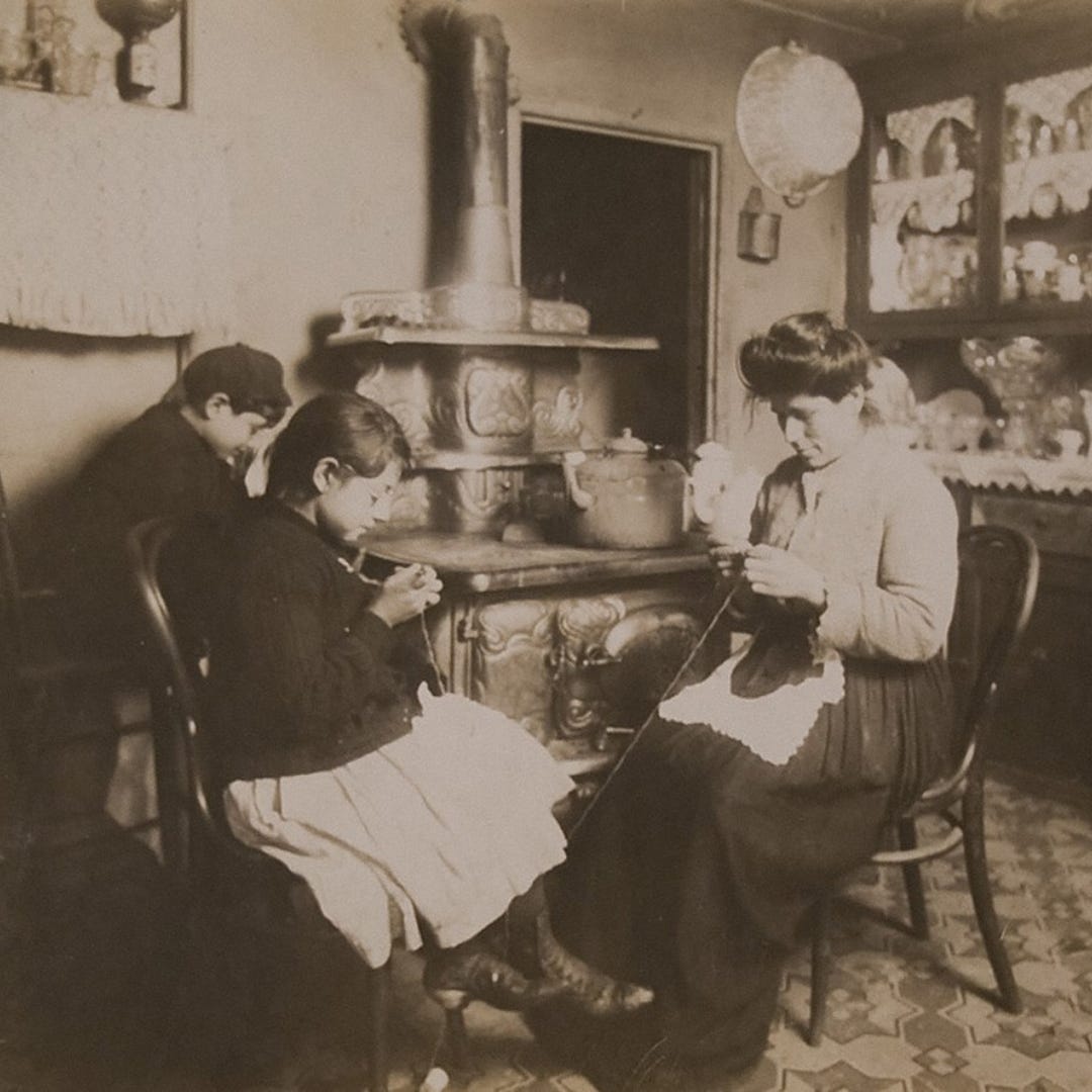 ce-making home work in New York City, 1912 CE. A mother and two children around age 10 to 13 sit around a stove and stitch lace in a NYC small tenement apartment. Behind the mother is a classic hutch filled with dish and glassware. In classic Italian-American style, a lace or dollie spread covers the hutch’s open display space. 