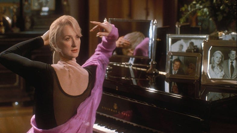 Film still from Death Becomes Her. Madeline's neck is twisted all around so that her head is looking behind her back. She's sitting at a piano.