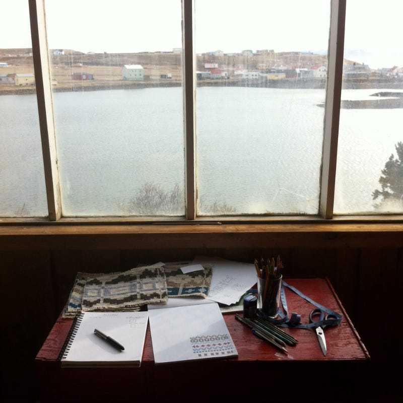 Desk with woven samples, notebook, pencils, scissors in front of a window looking out over a river