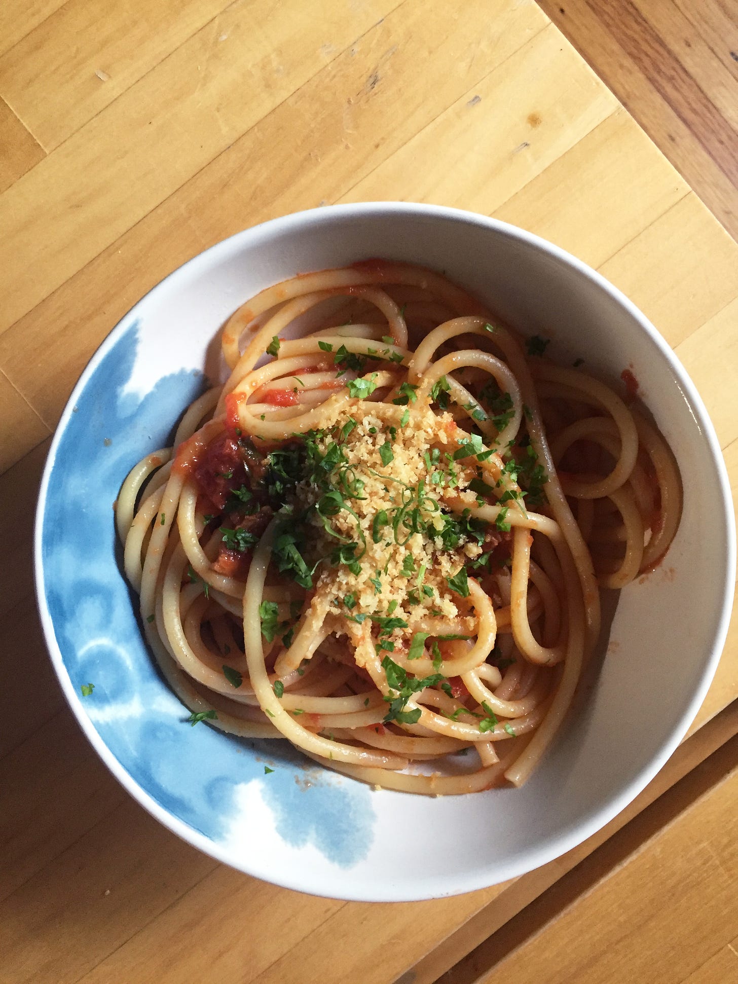 A white and blow bowl full of bucatini in red sauce and topped with basil, parsley, and toasted breadcrumbs.