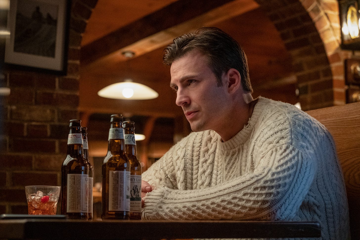 Chris Evans in a white Aran sweater sitting with his hands folded at a restaurant booth in KNIVES OUT. Four beer bottles and a half-drunk cocktail are also on the table.