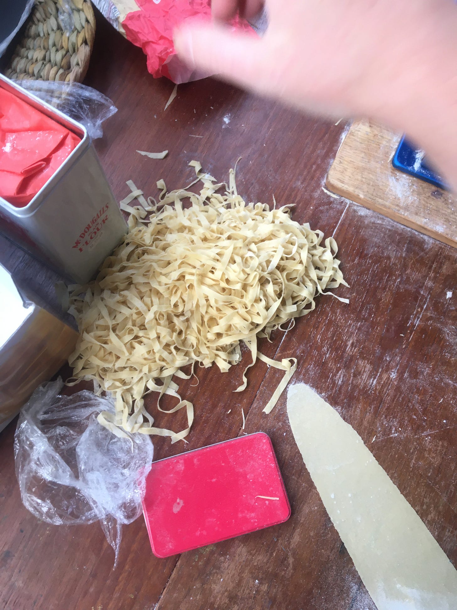 The same wooden table, with a pile of spangled pasta in ribbons. A smooth tongue shape of uncut pasta is to the bottom right, flat against the table. A tin of flour is to the left, its red lid too, a clod of cling film and some rush table mats. Kate's hand is, blurry, above. 