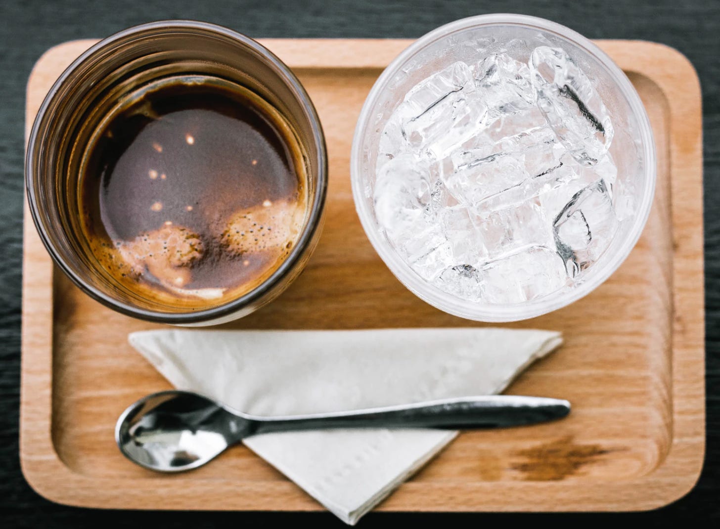 Top view of an espresso on the left and a cup of ice on the right and a silver stirring spoon on a white napkin at the bottom of the image. They sit on a blonde wood tray.