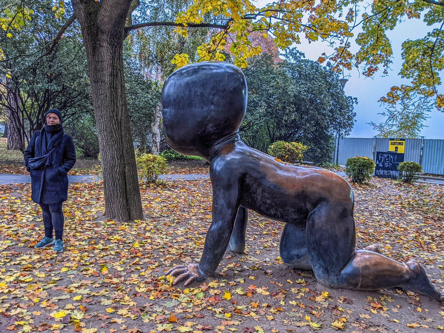 A woman stands next to a sculpture of a giant crawling baby in a park.