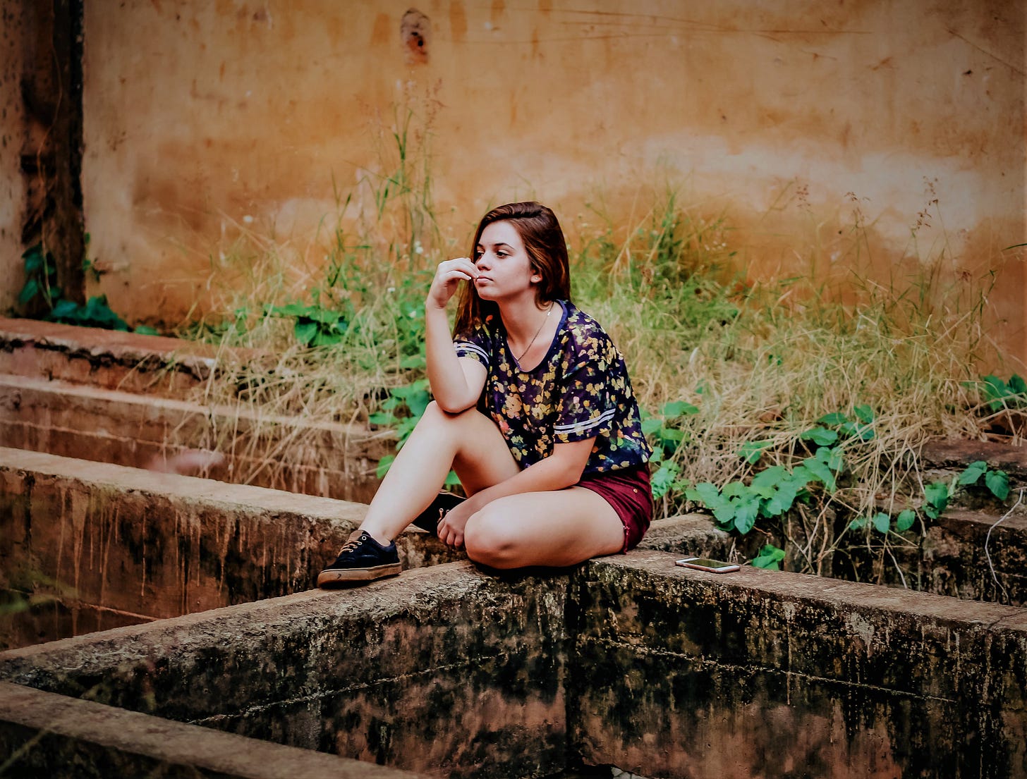 girl with brown hair wearing flowered top sitting on edge of concrete wall with hand to her cheek girl with brown hair wearing flowered top sitting on edge of concrete wall with hand to her cheek