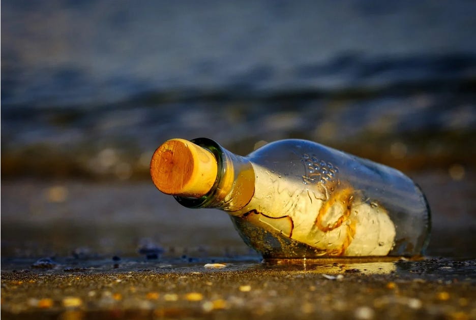 Foto de uma garrafa com uma cartinha dentro e uma rolha fechando o pote. Ela está meio atolada na areia, talvez esperando alguém encontrar, ou só cumprindo sua missão de levar a mensagem? 