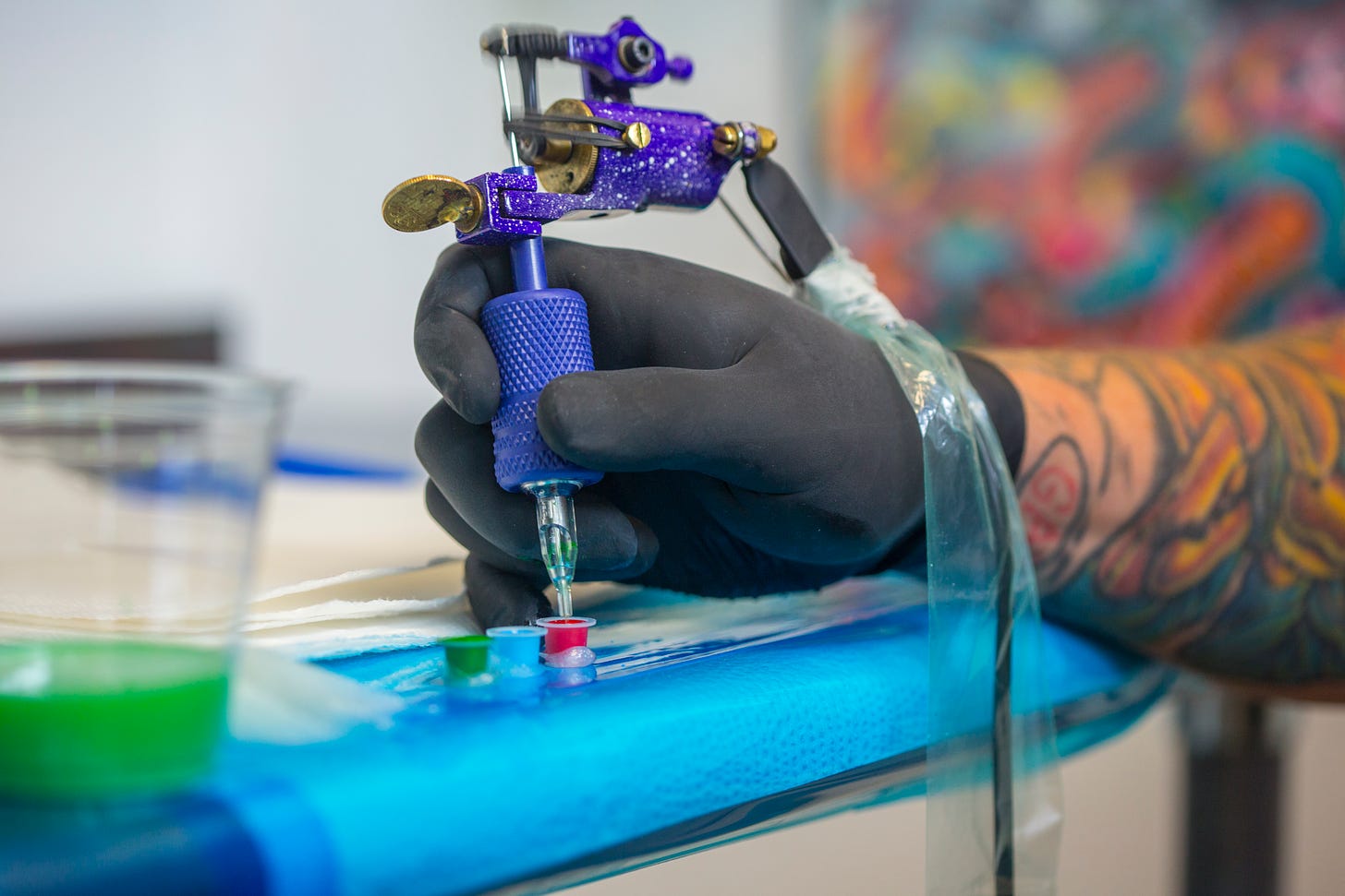 A black gloved tattooed arm dipping a blew modern tattoo machine in red ink