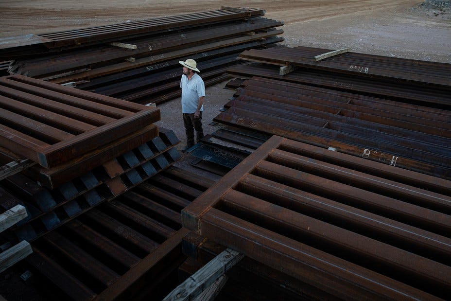 A man (Myles Traphagen, the borderlands program coordinator at the Wildlands Network) stands surrounded by piles of abandoned steel border wall sections in Arizona.