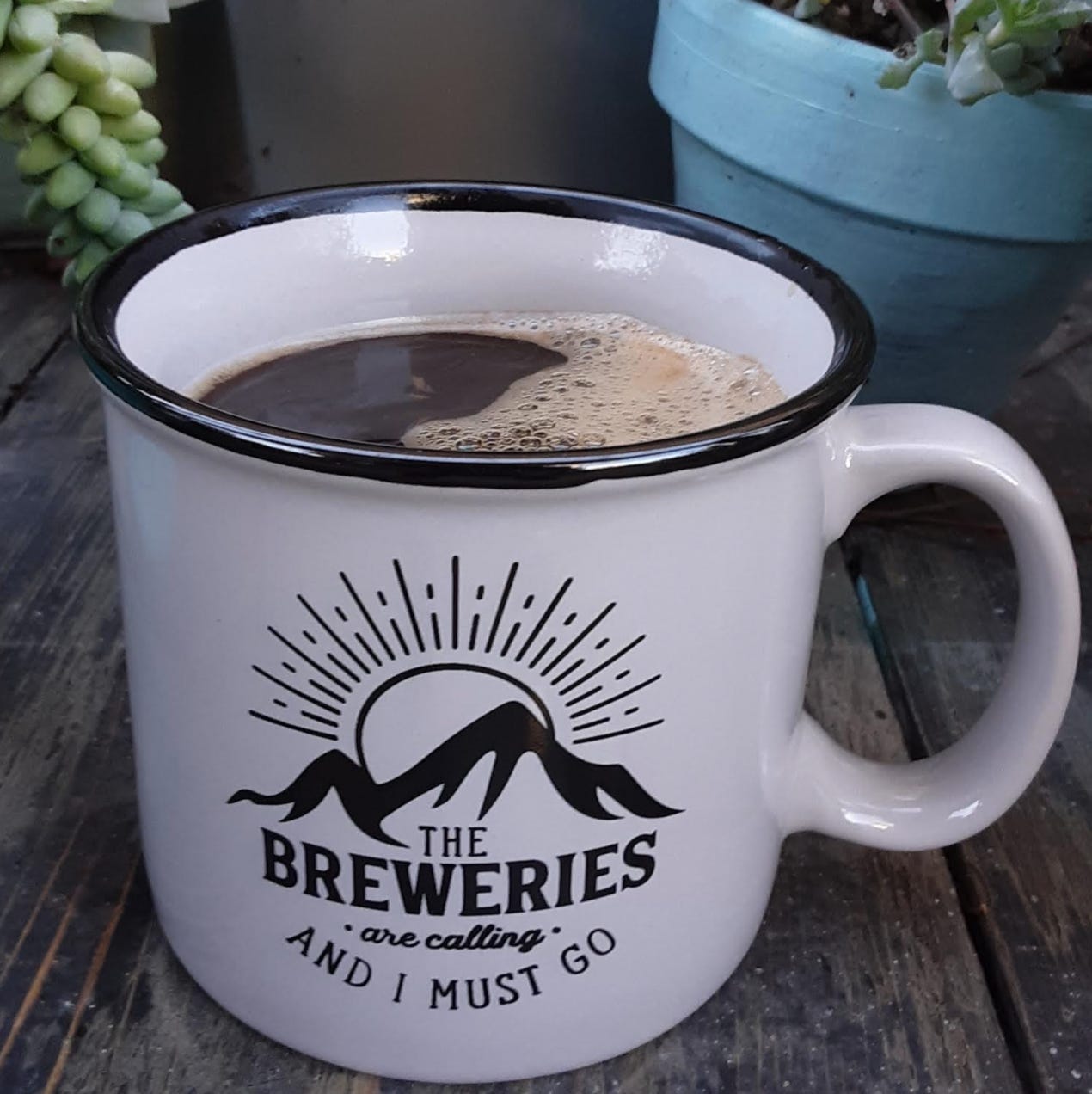 A white ceramic coffee mug with a black silhouette of a mountain and sunset behind it. Type saying, "The Breweries Are Calling and I must Go!" is on it. It is filled with coffee with blue planters filled with succulents in the background.