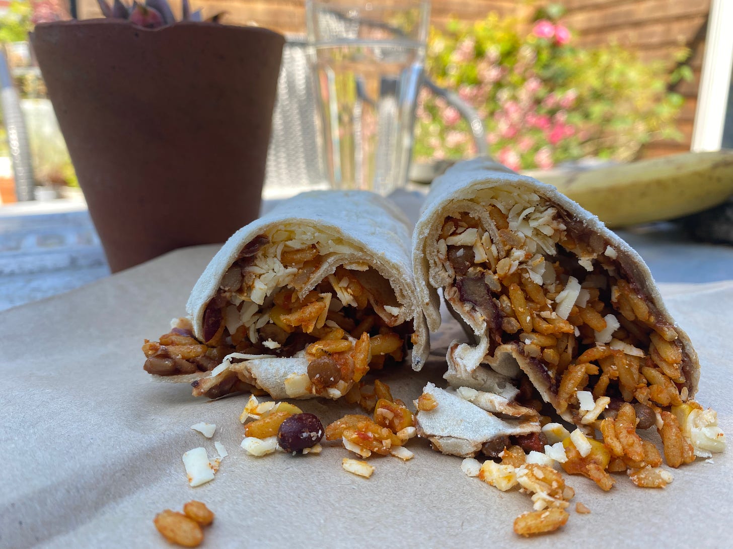 Sliced open burrito with its filling falling out, showing rice, beans and cheese. A terracotta pot, banana and glass of water in the background.