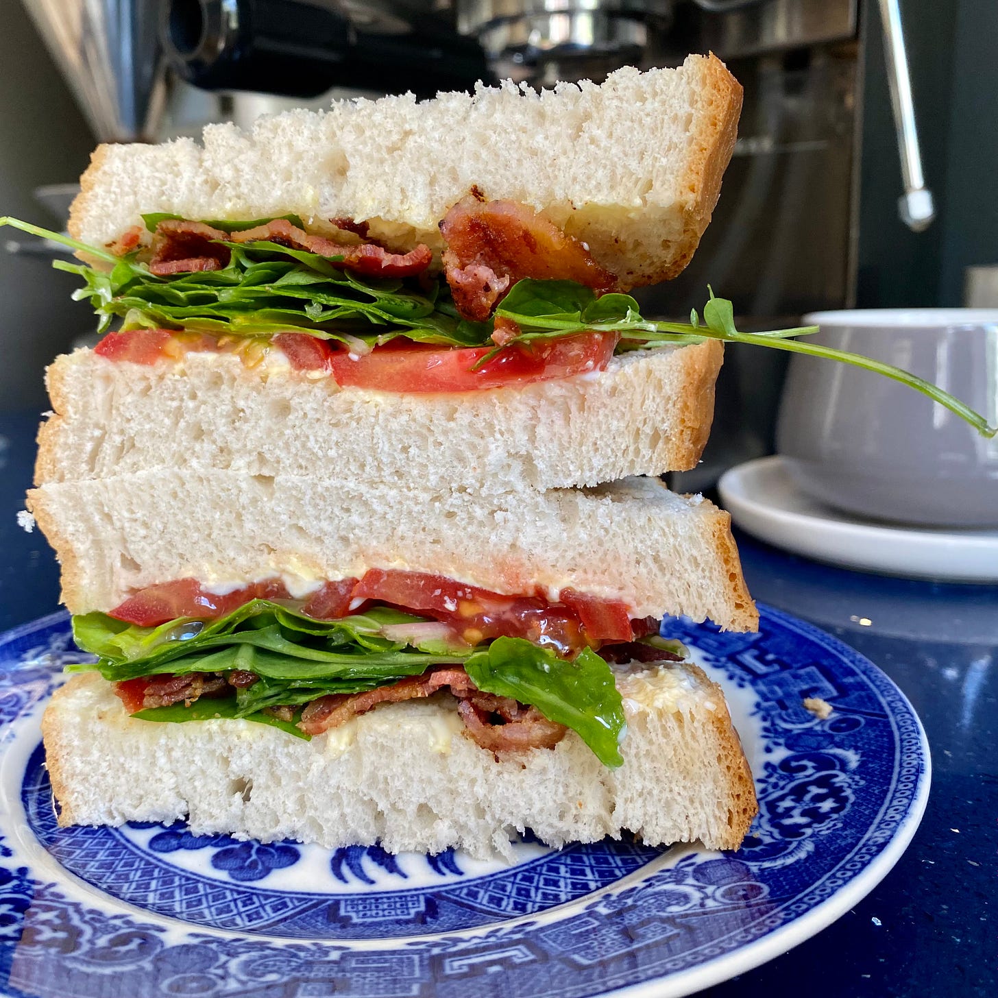 Cross-section of a sandwich: white bread filled with sliced tomato, bacon and salad leaves. Sandwich is on a blue and white patterned plate. A grey coffee cup is behind and to the right.