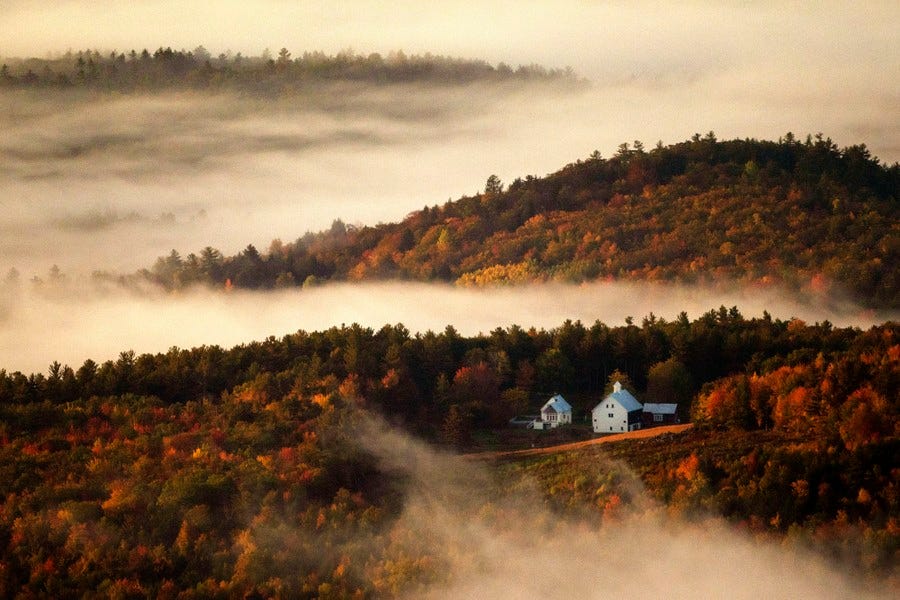 Fog flows in valleys between hills covered in fall-colored trees. A farm sits in the foreground.