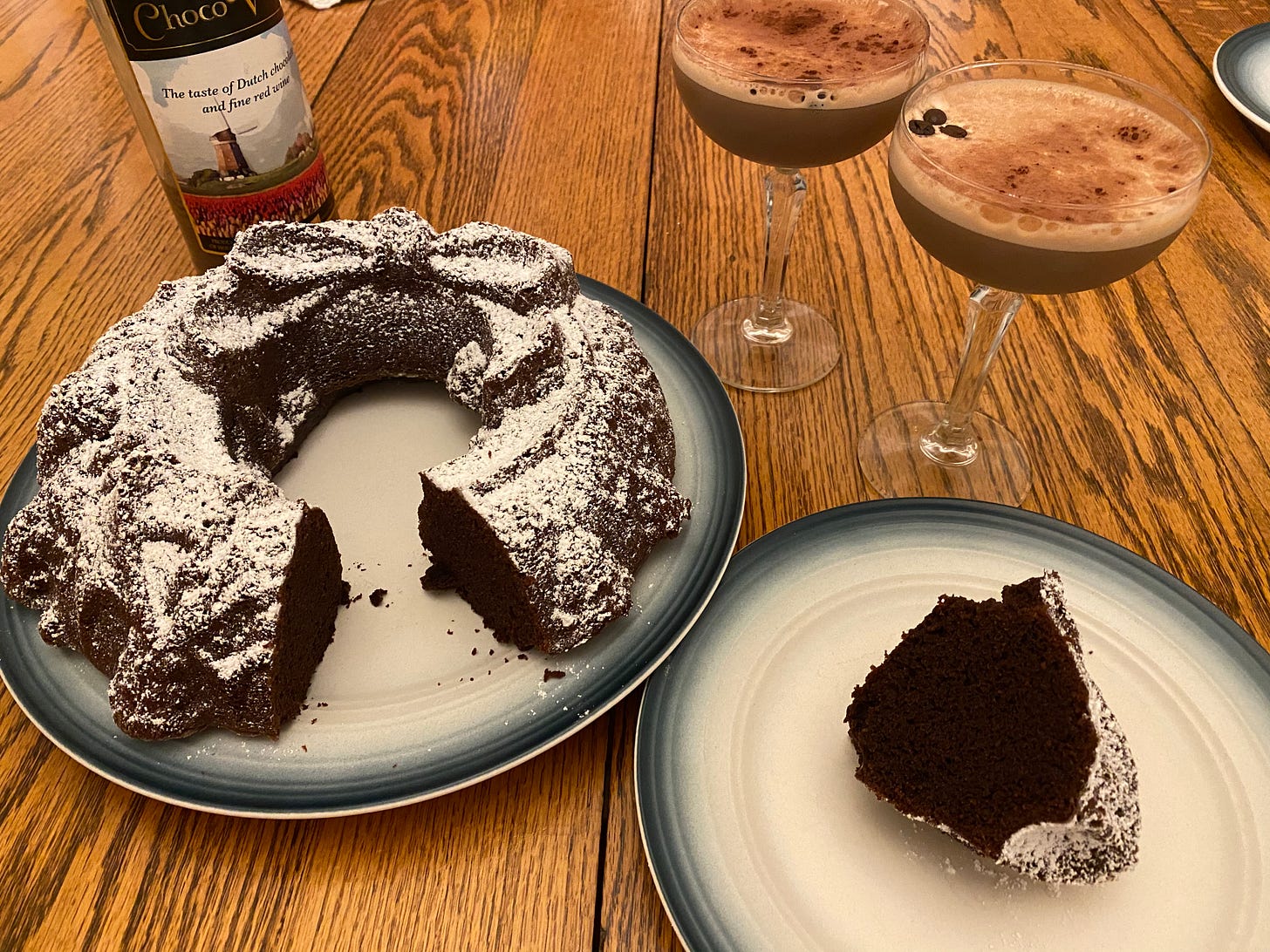 A deep brown chocolate bundt cake, dusted with powdered sugar, with one slice removed and plated nearby. Two espresso martinis in coupe glasses are in the background. 