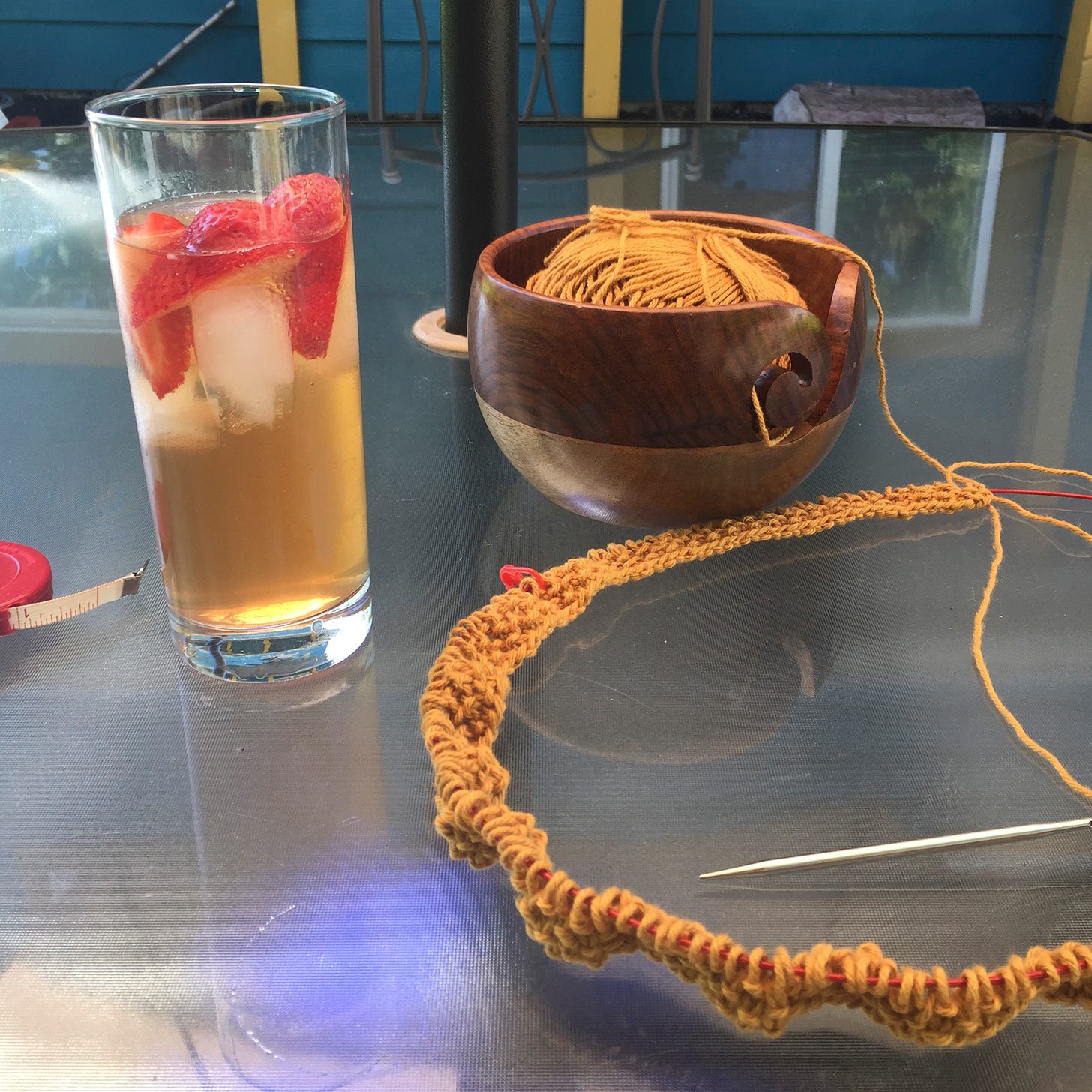 On an outdoor table is a Collins glass of iced tea with ice cubes and pieces of strawberry. Next to it is a two-toned wooden yarn bowl and a few rows of mustard yellow knitting on long circular needles.