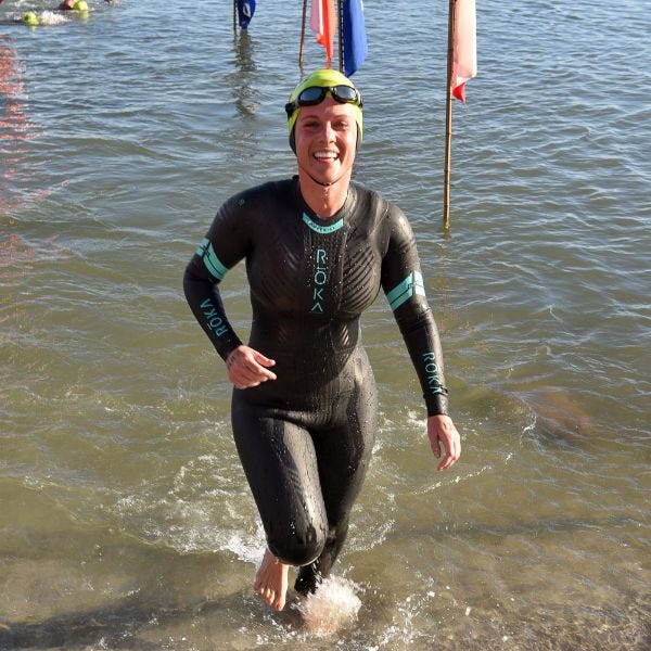 a mid-20s white woman in a wetsuit, swim cap, and goggles jogging out of the ocean and smiling