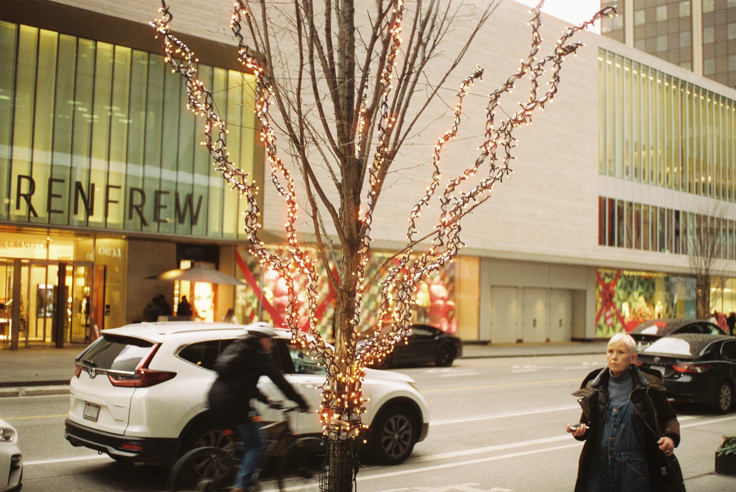 A photograph of a tree with Christmas lights on a busy street, and in the foreground right side a woman is walking down the sidewalk.