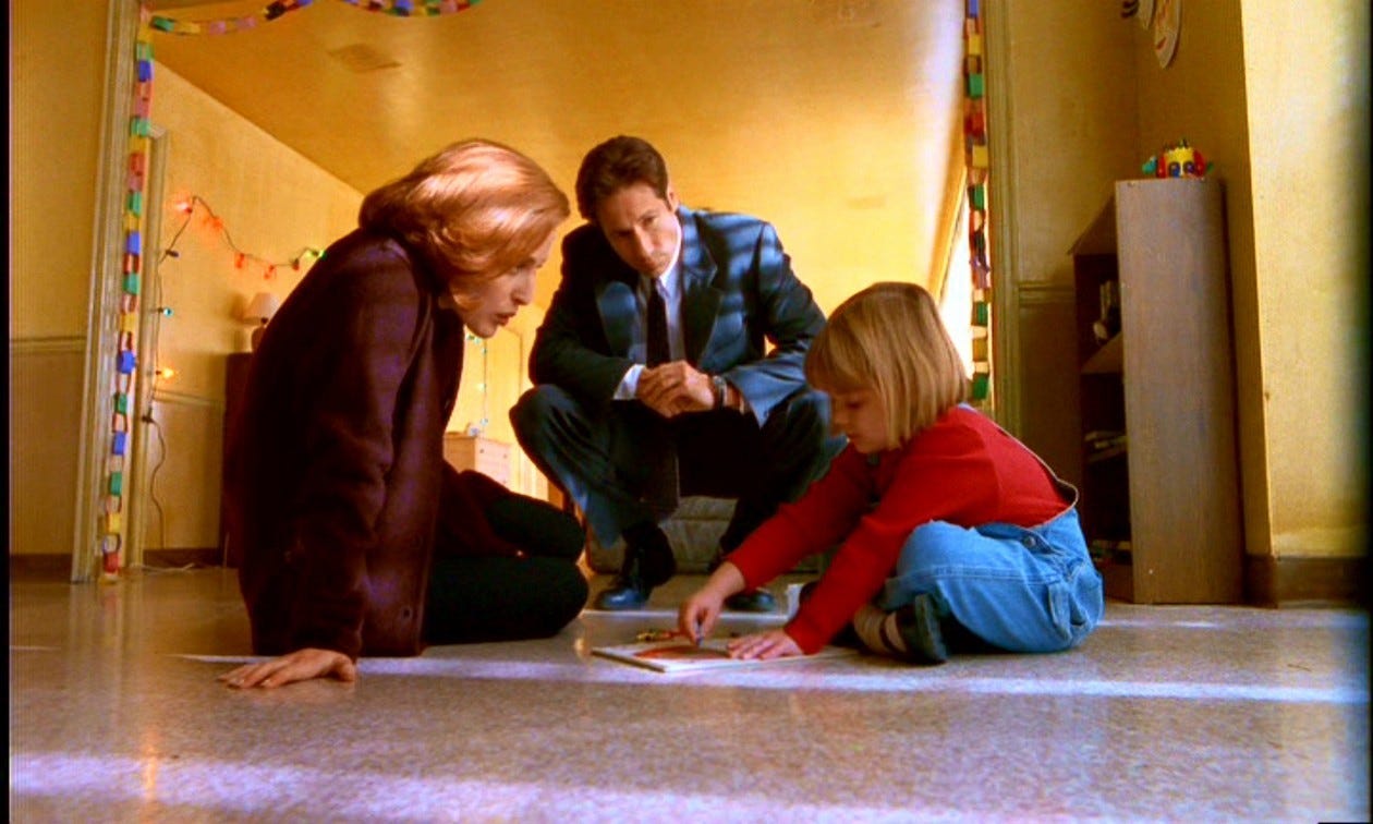 Scully sitting on the floor next to a child who's playing, looking interested and supportive; Mulder is perched on a stool looking concerned.