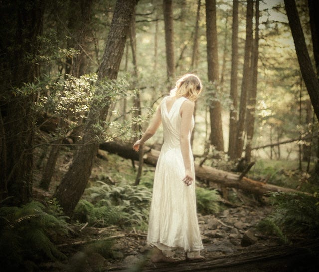 A blond haired white woman in a romantic white silk dress barefoot in a sunlit forest 