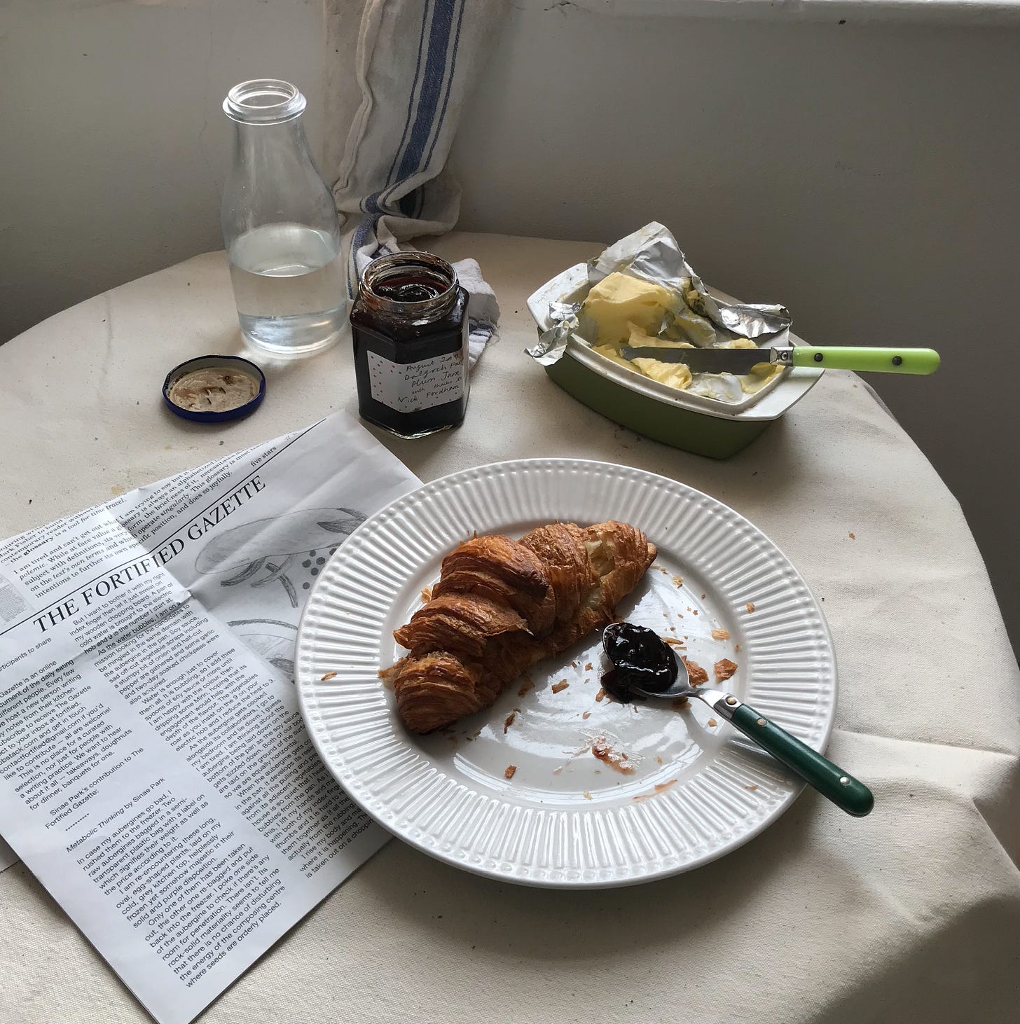 The same round table, set, but earlier, with the croissant intact, and a spoon, full of jam, beside on the plate. In the butter dish, a knife is askew.