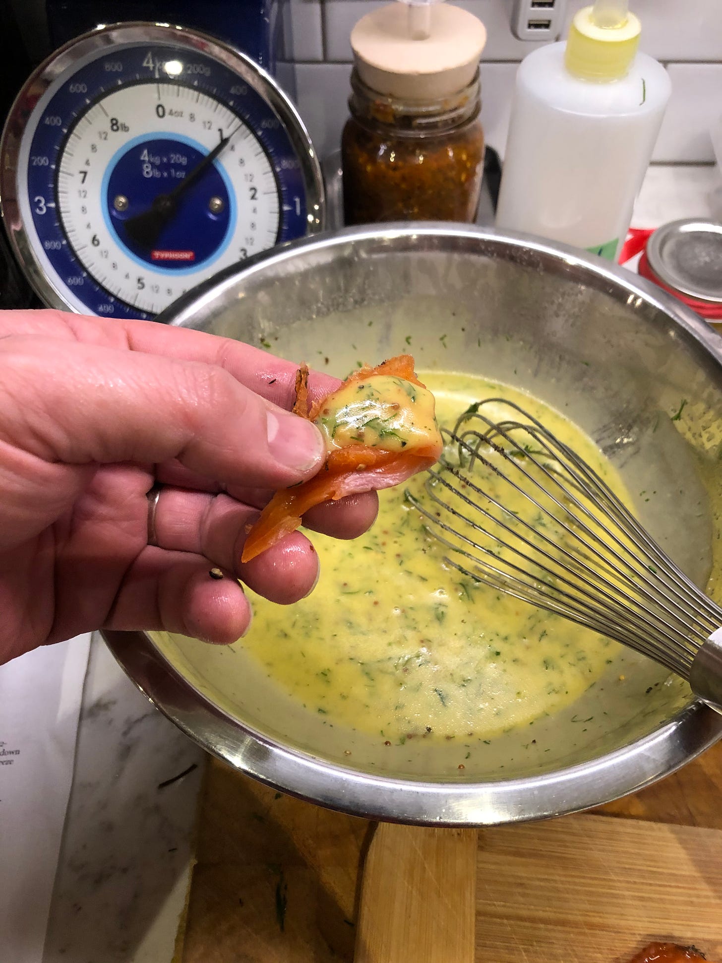 A thin slice of gravlax with a small dollop of mustard-dill sauce, being held over a mixing bowl of just-made sauce.