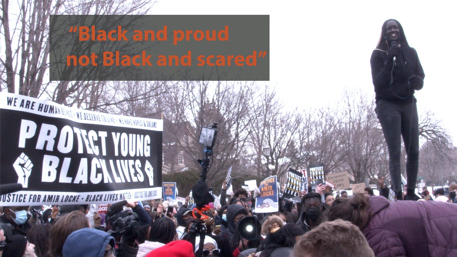 young Black girl stands above crowd next to a sign reading "protect young black lives"