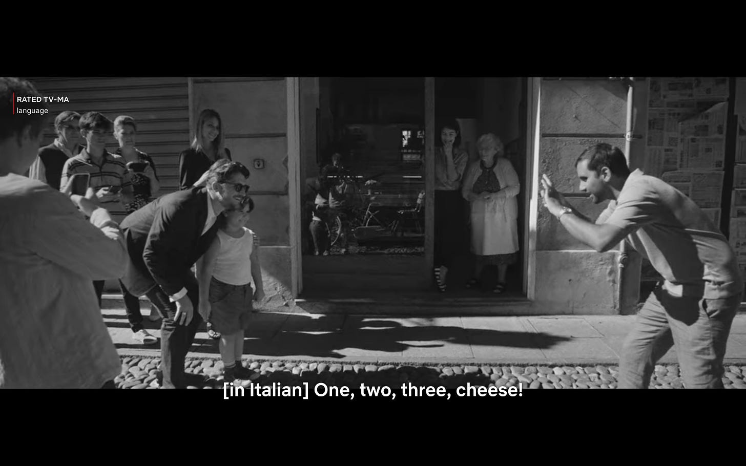 Master of None scene: Black and white film, Dev small Indian man taking photo of young Italian girl and famous Italian soccer player. The subtitle says "[in Italian] One, two three cheese!