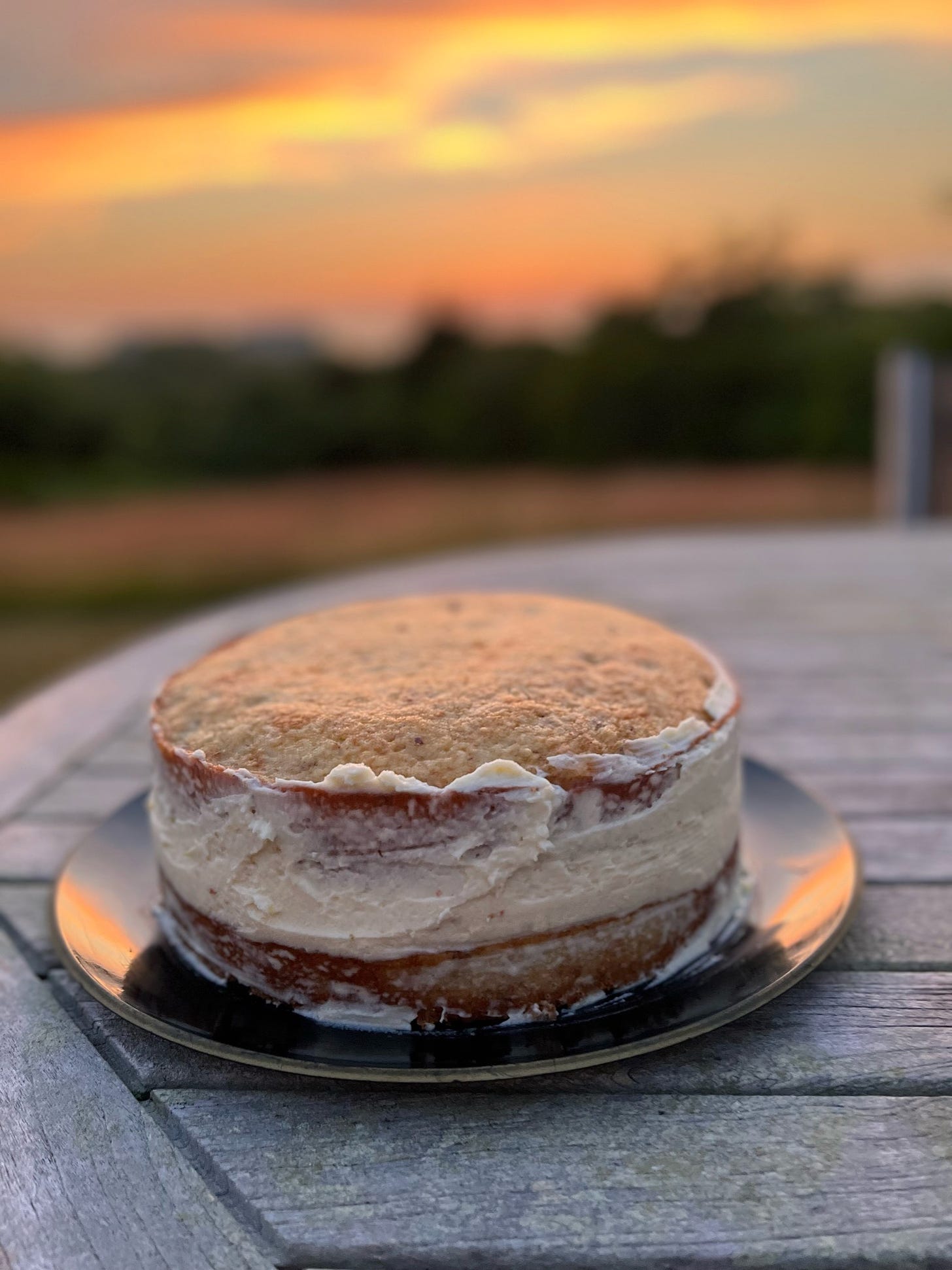A two layer, semi-frosted almond cake sits on an outdoor table at sunset.