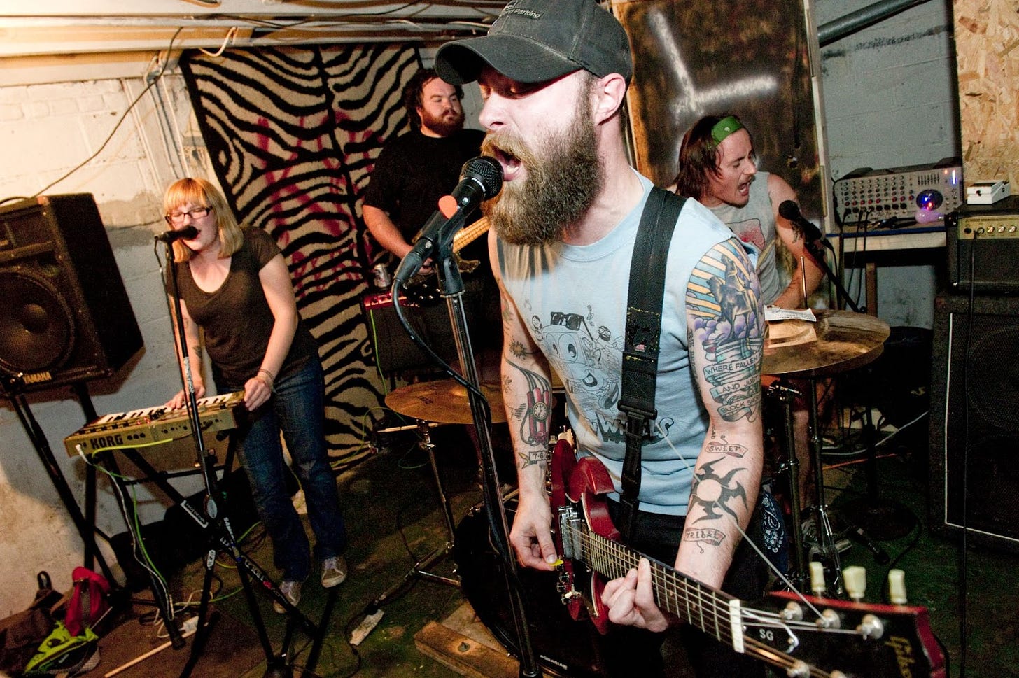 From left to right, Adrienne is standing in a brown top and jeans singing into a microphone and playing keyboard; Richard is on bass in a black tshirt and cool beard; Louis is in the back right on drums and vocals, and my husband Matt is in the front in a gray tshirt and hat, cool tattoos, and a red electric guitar while he sings into a microphone. 