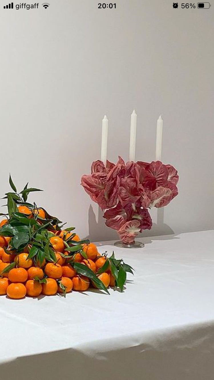 On a white tablecloth, a pile of tangerines with their stems and leaves, stacked in a pyramid shape. To the right a candelabra made of pink cabbage leaves, with three white candles. 